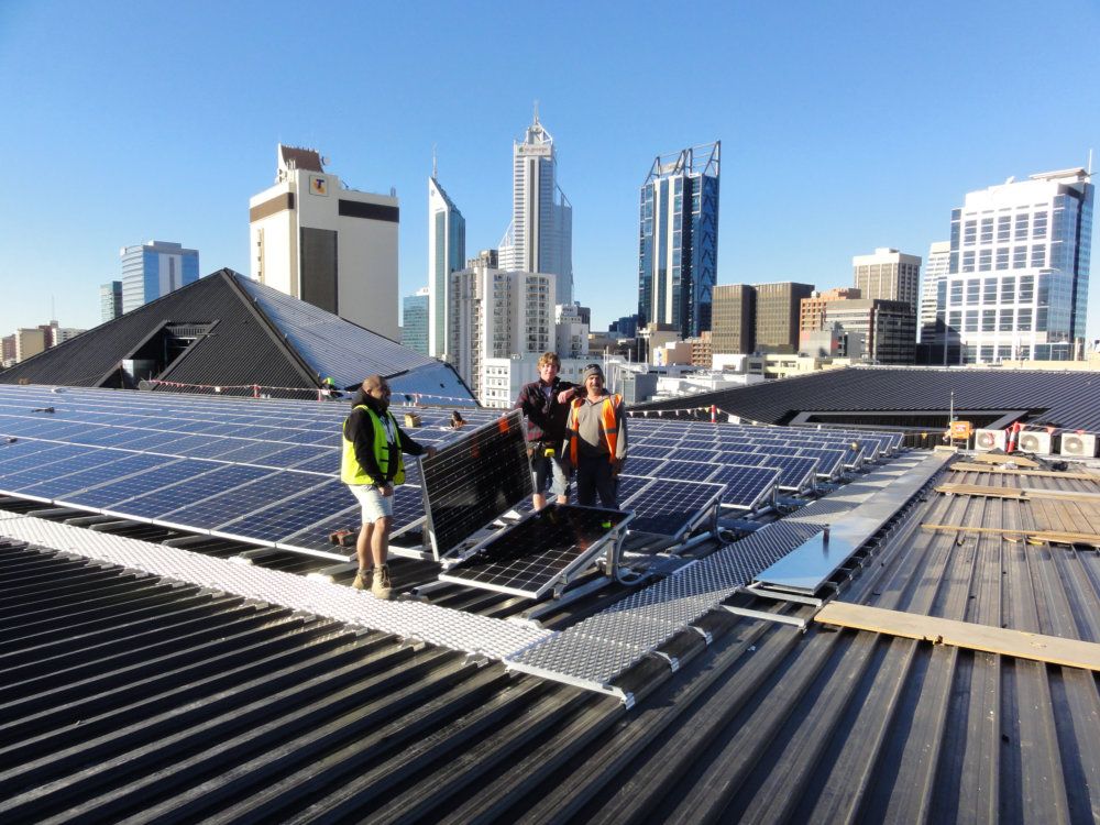 Installers with solar panels on the Perth Arena roof, city towers behind